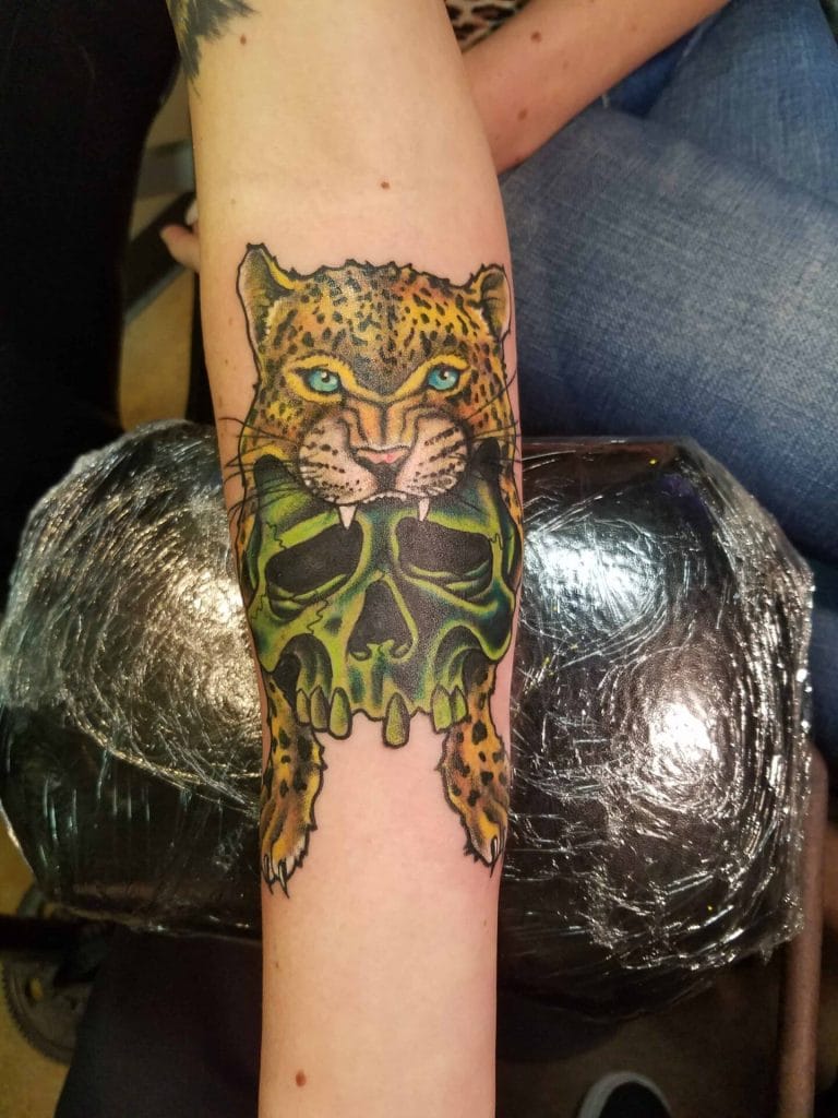 Leopard face with blue eyes above a green skull tattoo on a persons forearm.