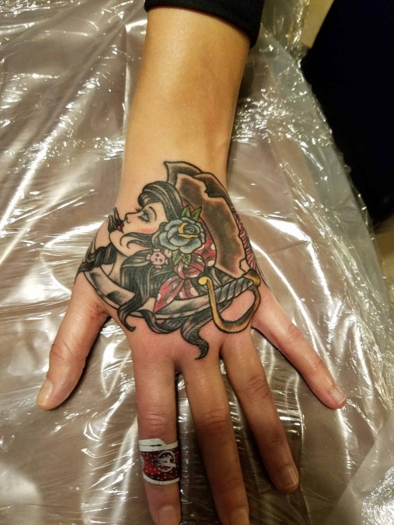 Tattoo of a woman with long black hair, flowers, and a hat on the hand.