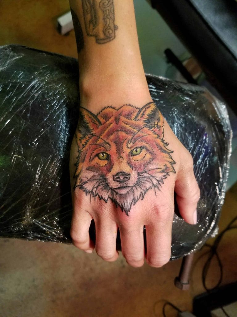 Colorful fox face tattoo on hand with yellow eyes and detailed fur, wrist tattoo visible.