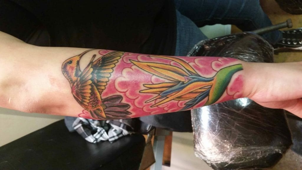 Colorful hummingbird and flower tattoo on forearm with pink clouds background.
