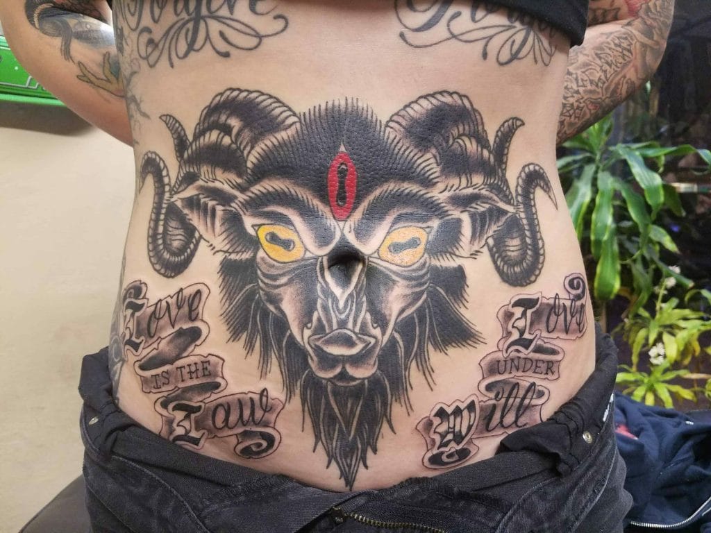 Tattoo of a goats face with yellow eyes, red mark on forehead, and text Love is the law, love under will on the stomach.