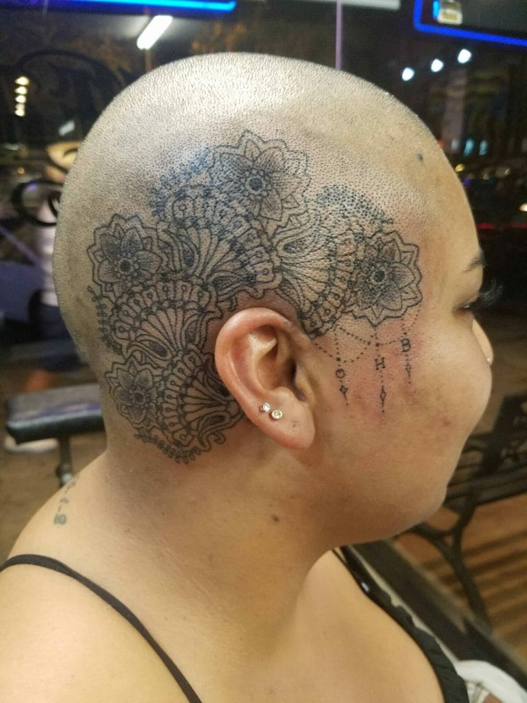 Person with a shaved head, floral tattoo on scalp, and small earrings, sitting indoors.