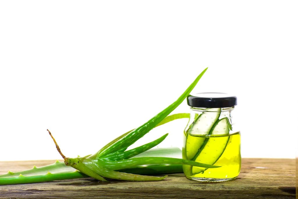 Aloe vera leaf and a small glass bottle of yellow liquid with aloe slices, placed on a wooden surface against a white background—ideal for soothing tattoo sensitivities or calming mild tattoo reactions.