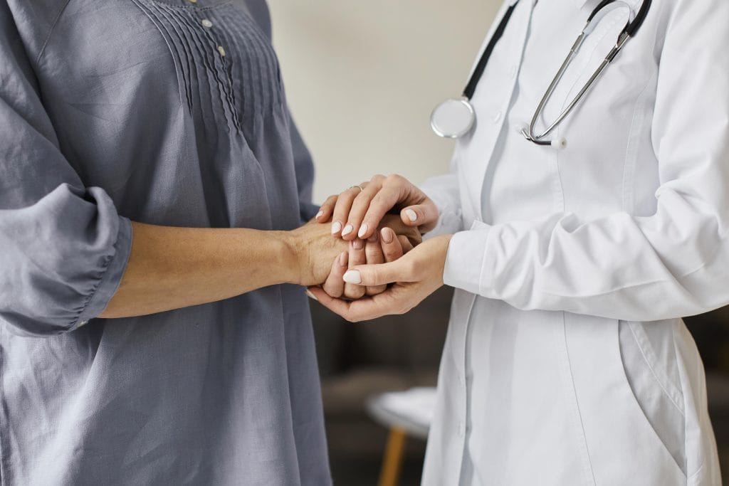 A doctor in a white coat holds the hands of a patient in a blue shirt, suggesting support or a consultation about medical procedures in a healthcare setting.