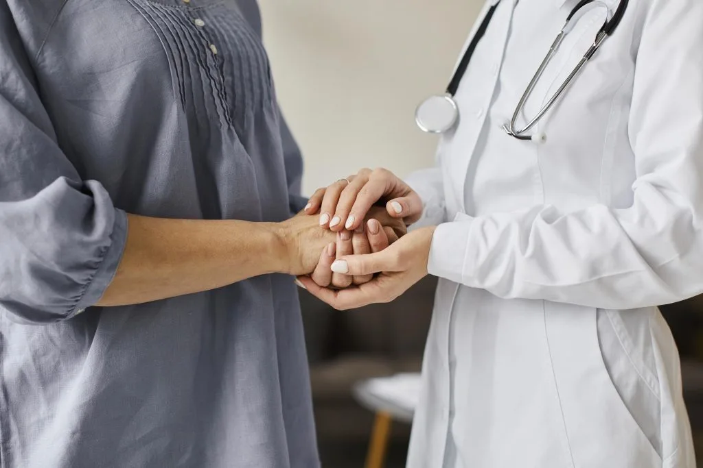 A doctor in a white coat holds the hands of a patient in a blue shirt, suggesting support or a consultation about medical procedures in a healthcare setting.