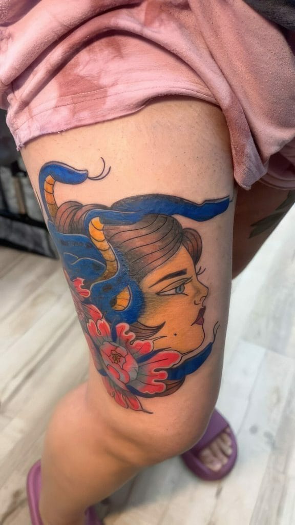 Tattoo of a womans face with flowing hair and a blue snake intertwined, surrounded by red flowers.