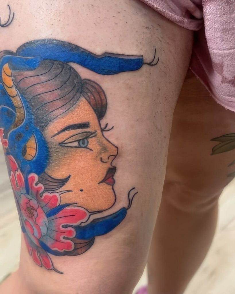 Tattoo of a womans face with blue hair and pink flowers on upper thigh.