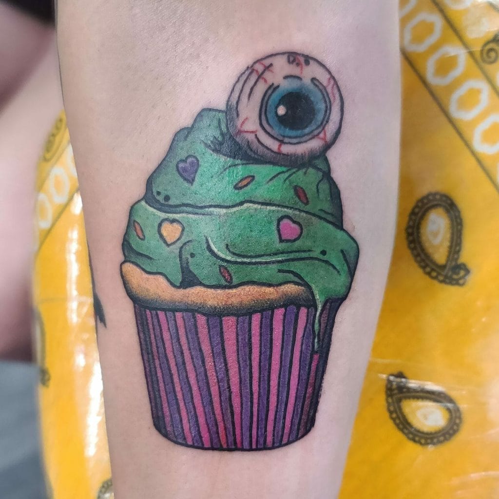 Colorful cupcake tattoo with a green frosting, pink and purple hearts, and a large eyeball on top.