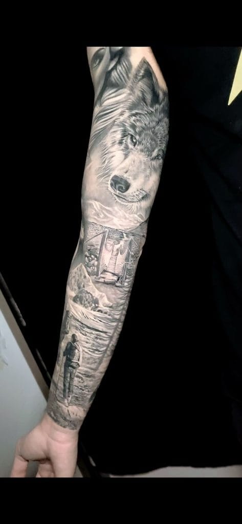 Detailed black and gray tattoo sleeve featuring a wolfs face, mountains, a bridge, and a person standing by a lake.