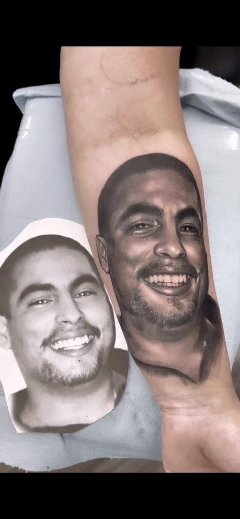 Realistic tattoo of a smiling mans face on a persons forearm, with a printed photo of the same face nearby.