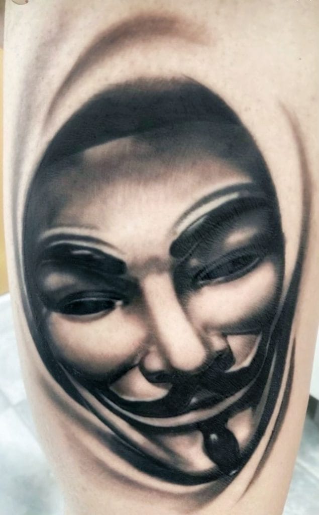 Tattoo of a smiling Guy Fawkes mask on persons arm in black and gray ink.