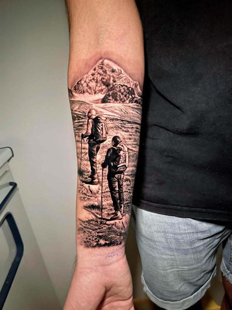Tattoo of two hikers walking in a mountainous landscape with snow-capped peaks in the background.