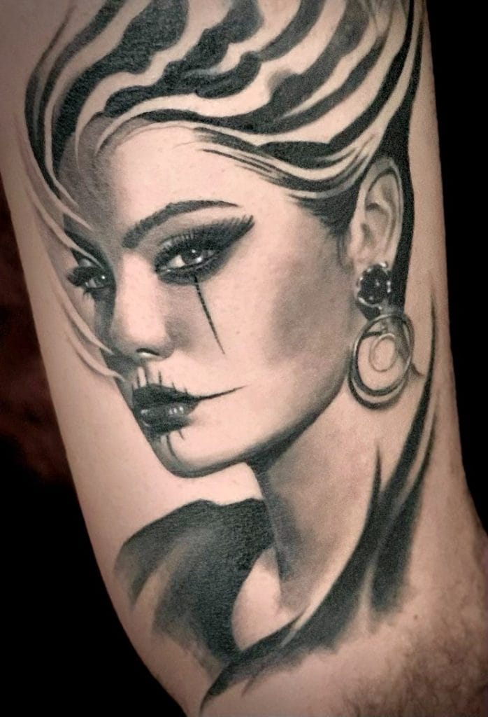Black and gray realistic tattoo of a woman with bold makeup, striped hair, and large earrings.