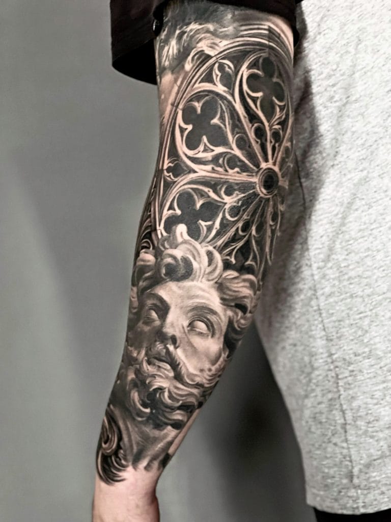 Detailed black and gray tattoo of a bearded mans face and ornate floral patterns on the arm.