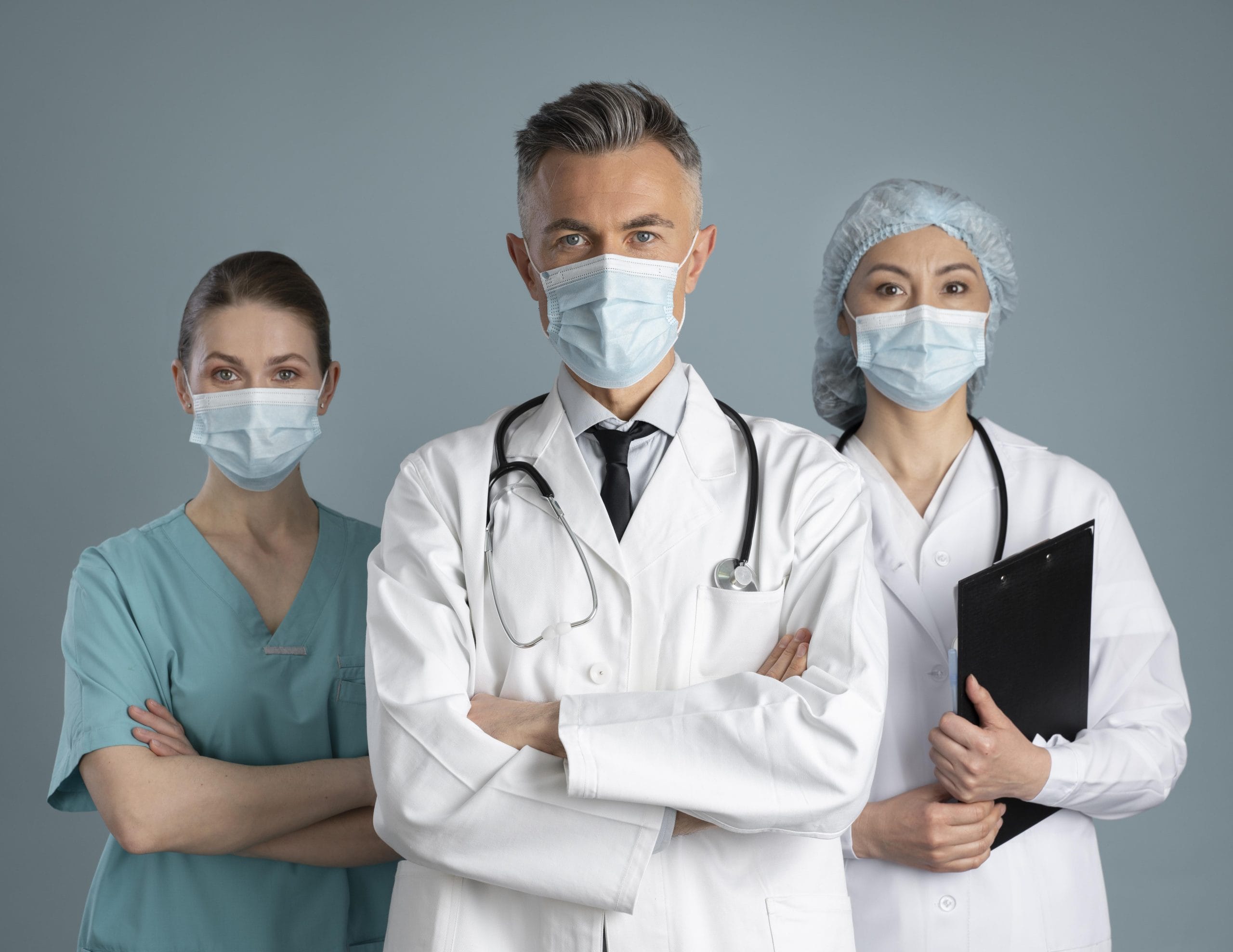 Three healthcare professionals—one doctor with a stethoscope—wear medical masks and uniforms as they stand together, ready for medical procedures; one holds a clipboard, showing teamwork in healthcare.