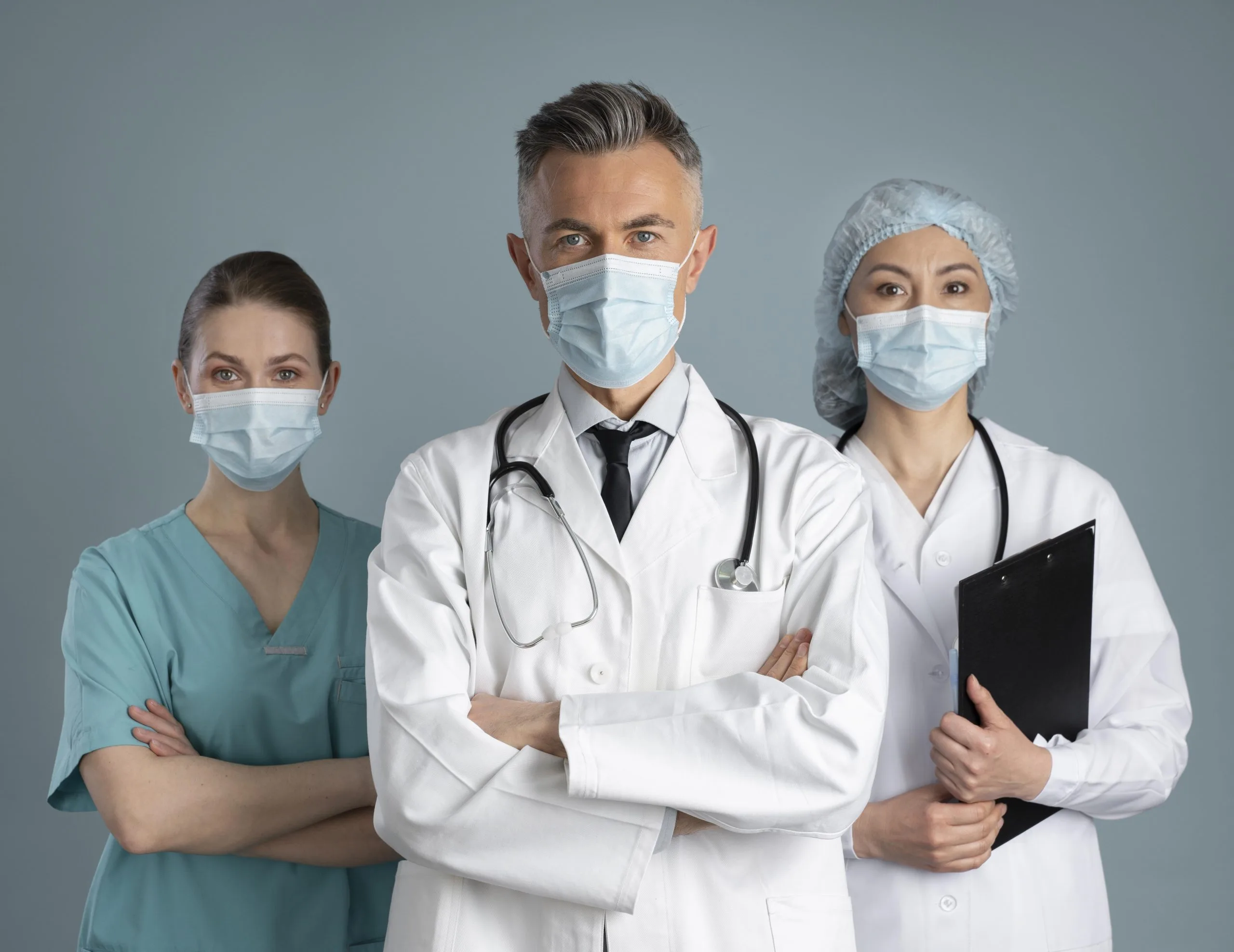 Three healthcare professionals—one doctor with a stethoscope—wear medical masks and uniforms as they stand together, ready for medical procedures; one holds a clipboard, showing teamwork in healthcare.
