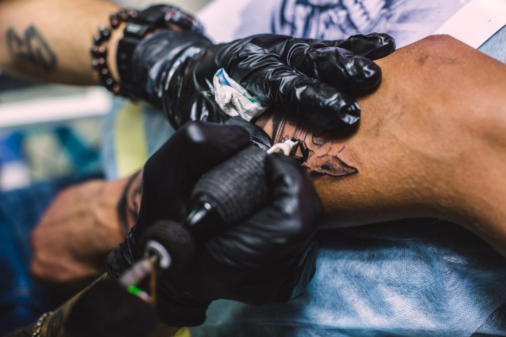 A tattoo artist wearing black gloves is using a tattoo machine to ink a design with white ink highlights on a person's forearm.