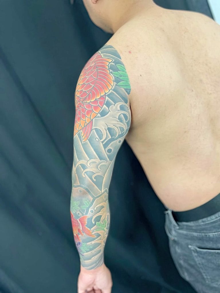Colorful koi fish and wave tattoo covering the persons upper arm and shoulder.