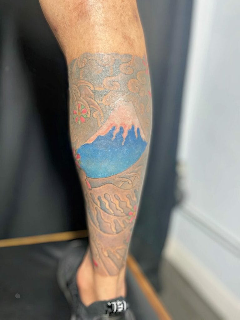 Tattoo of Mount Fuji with clouds, pink cherry blossoms, and blue sky on a persons leg.