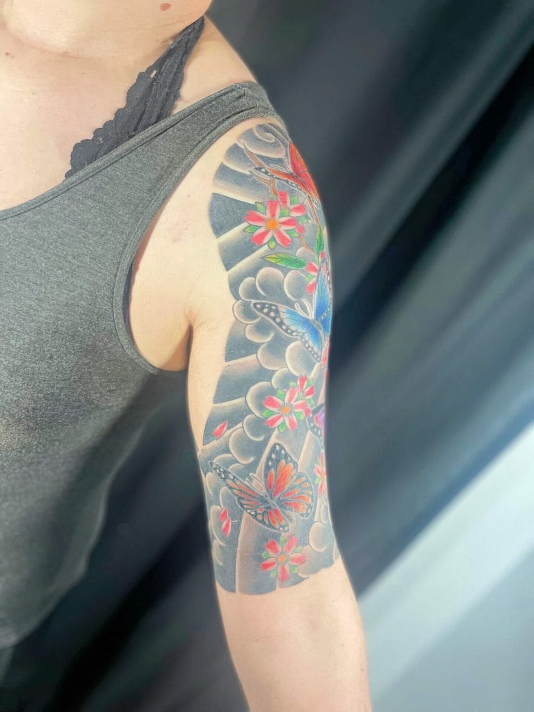 Colorful butterfly and flower tattoo on upper arm with clouds and shading details.