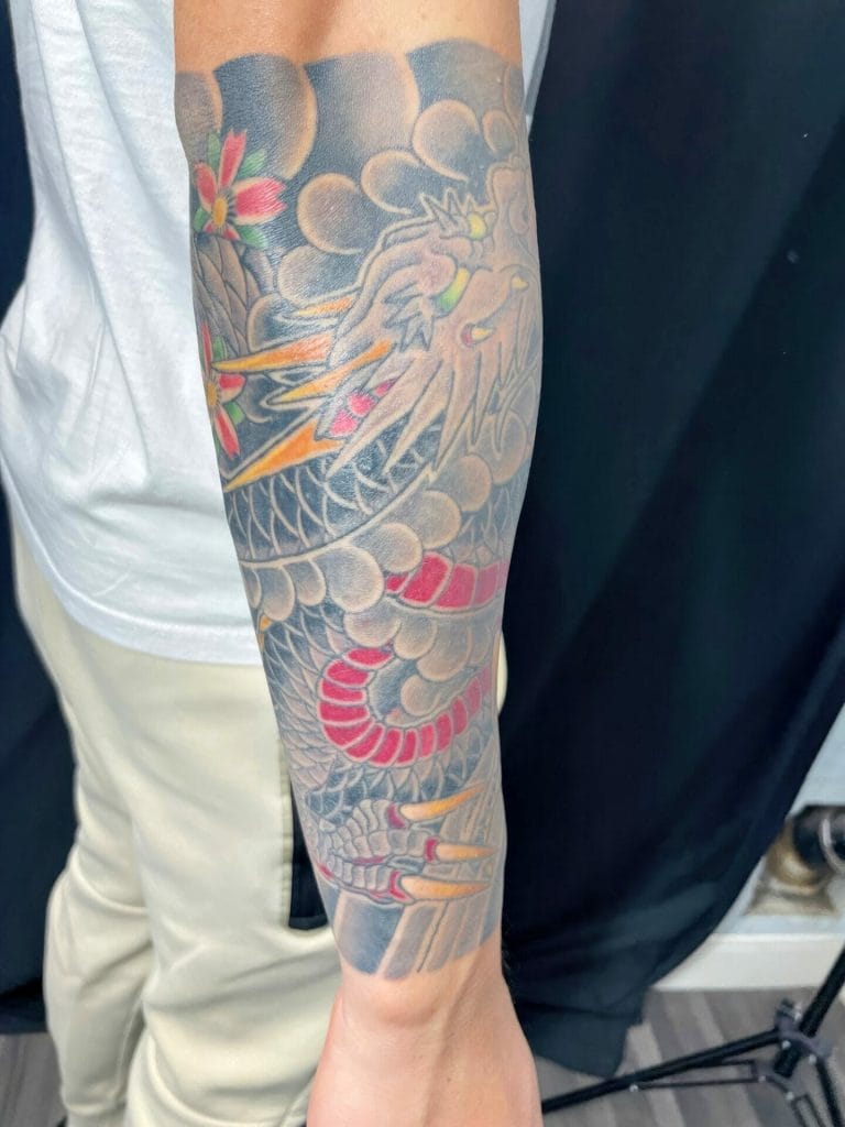 Colorful dragon tattoo with clouds, flowers, and flames on a persons forearm.