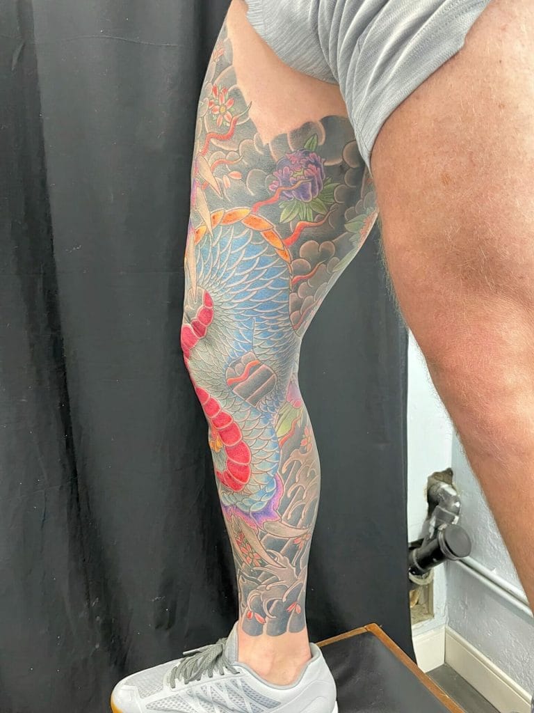 Colorful dragon tattoo covering the persons thigh and lower leg, with clouds and flowers.