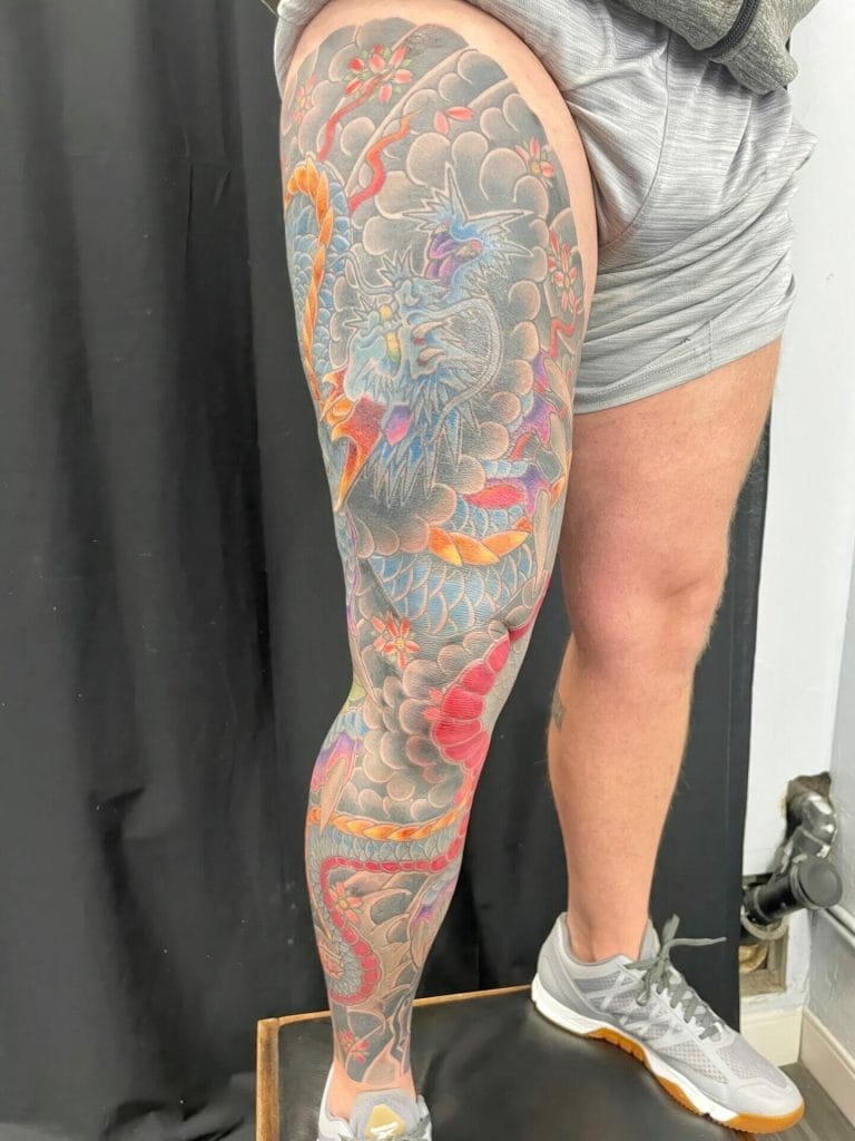 Colorful dragon tattoo covering the persons thigh and lower leg, with clouds and flowers.