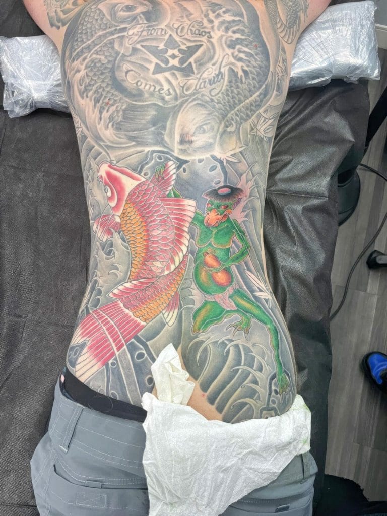 Colorful koi fish and a green frog tattooed on a persons back with a dragon and clouds in the background.