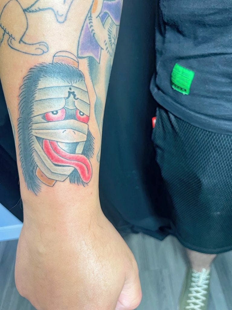 Tattoo of a cartoonish, smiling face with red eyes, a protruding tongue, and a furry outline.