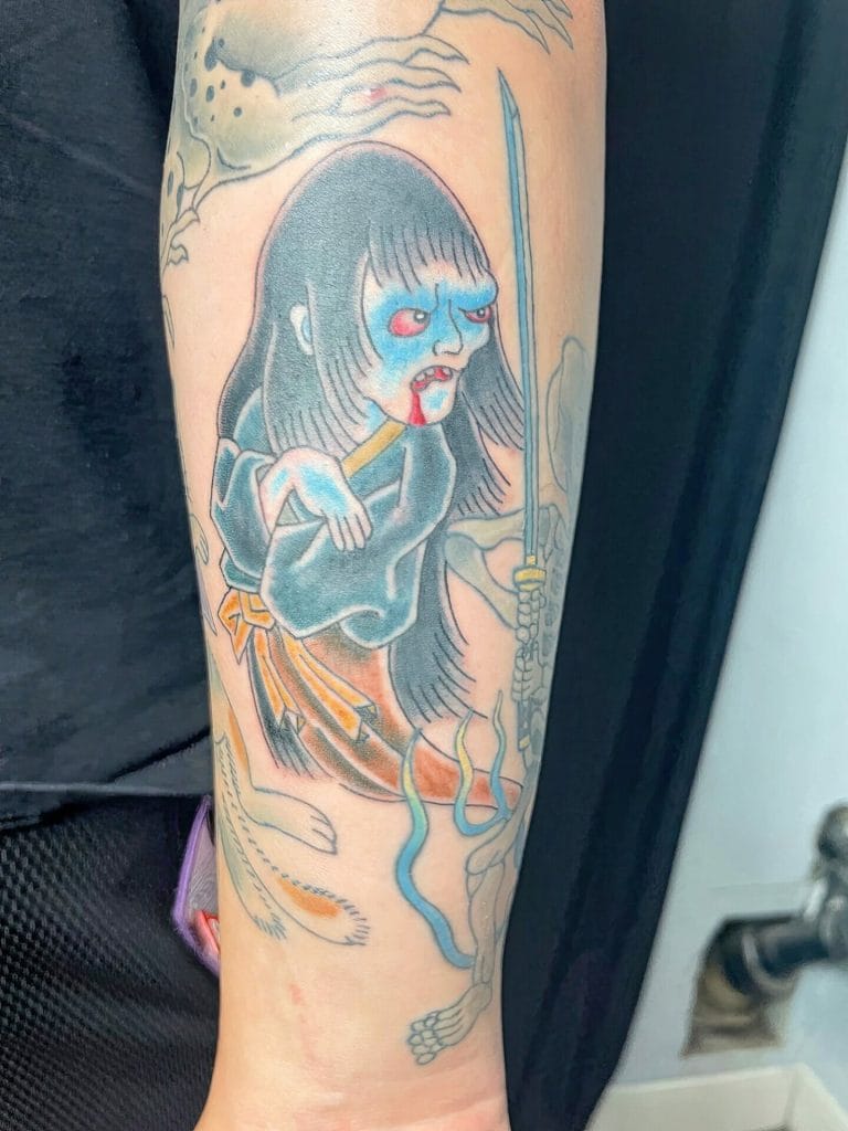 Tattoo of a fierce girl with blue face paint, long black hair, holding a sword, with a skeletal creature below.