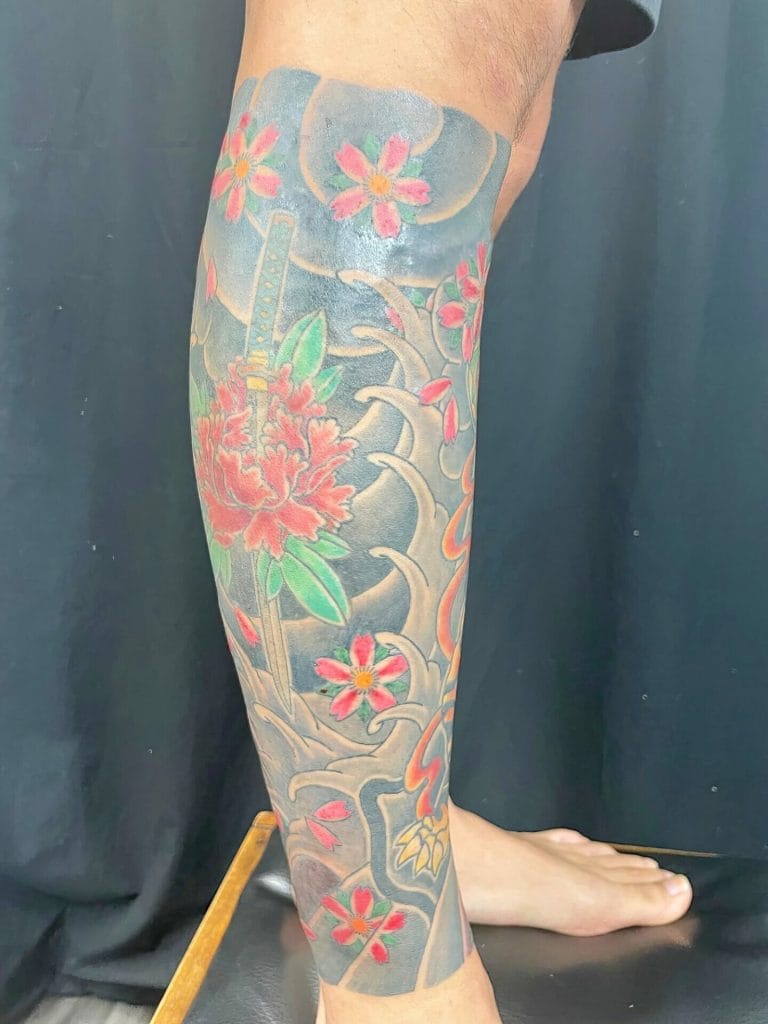 Colorful Japanese-style tattoo with flowers, a sword, and swirling clouds on a leg.