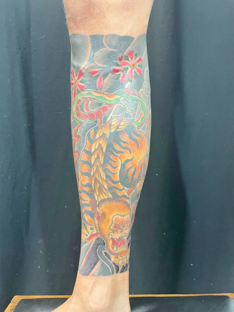 Colorful tattoo of a roaring lion, a snake, and cherry blossoms on a persons calf.