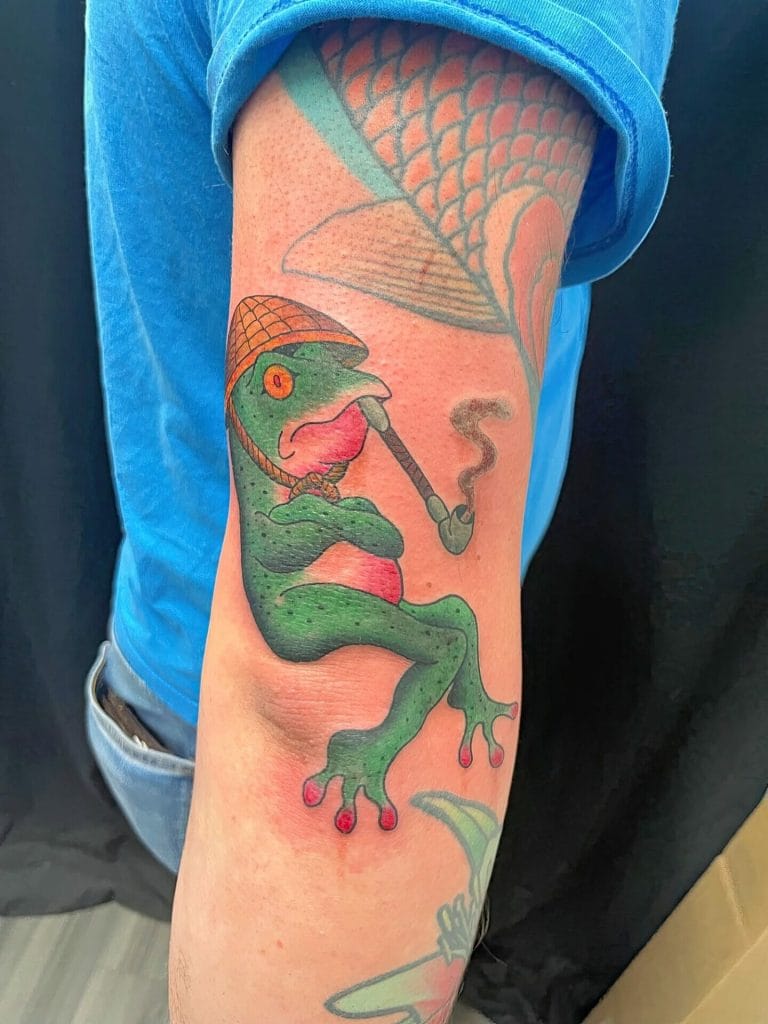 Green frog with orange eyes, smoking a pipe, wearing a hat, and sitting with crossed arms.