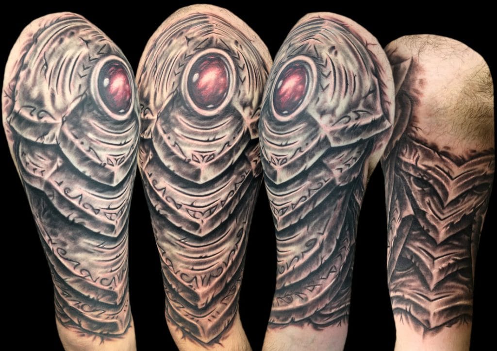 Detailed black and gray tattoo of a robotic eye and armor design on upper arm and shoulder.