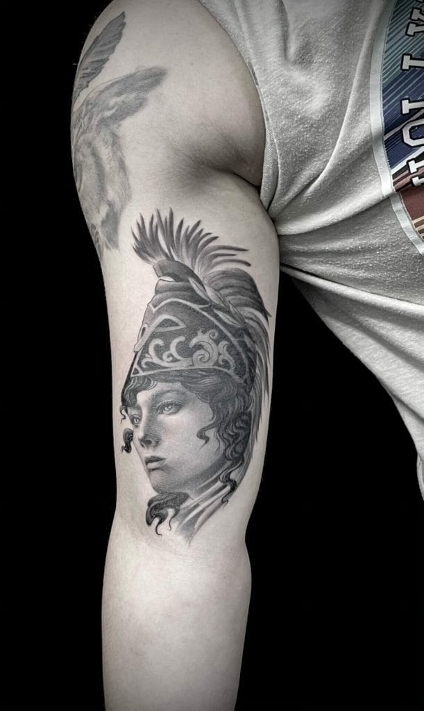 Black and gray tattoo of a womans face wearing a detailed helmet with a feathered crest on the upper arm.