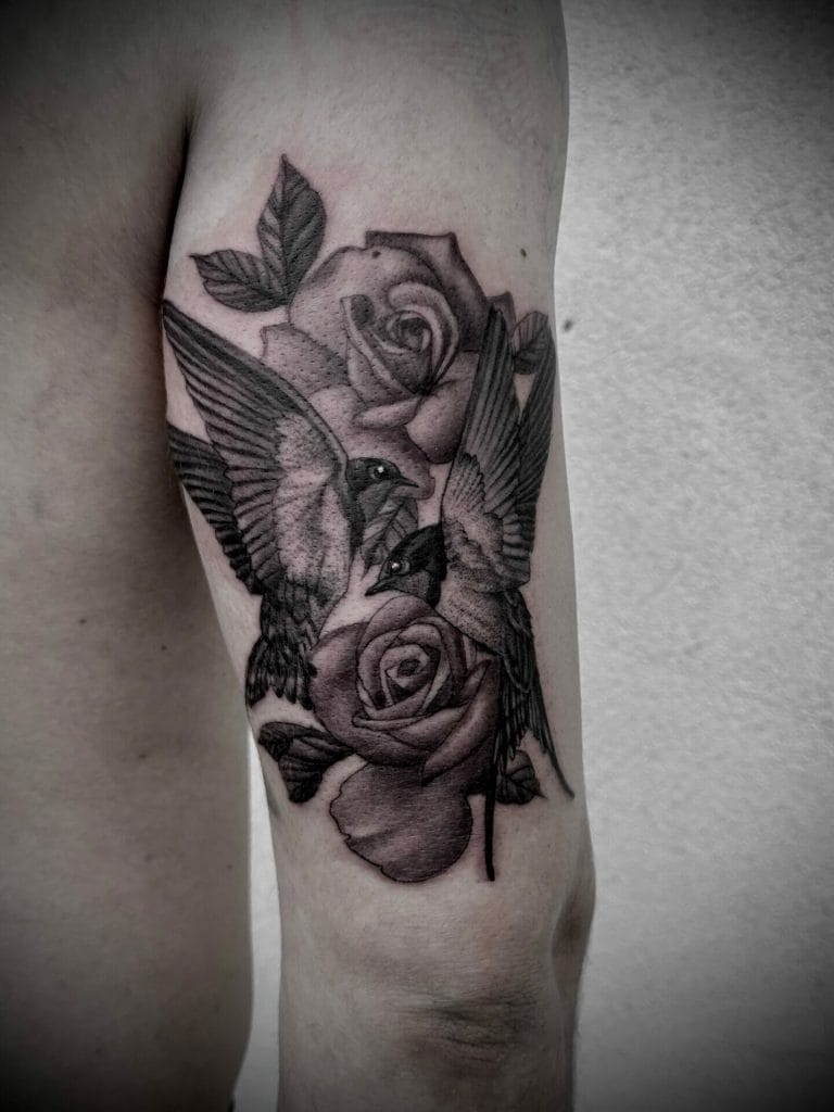 Black and gray tattoo of two hummingbirds and roses on a persons arm.