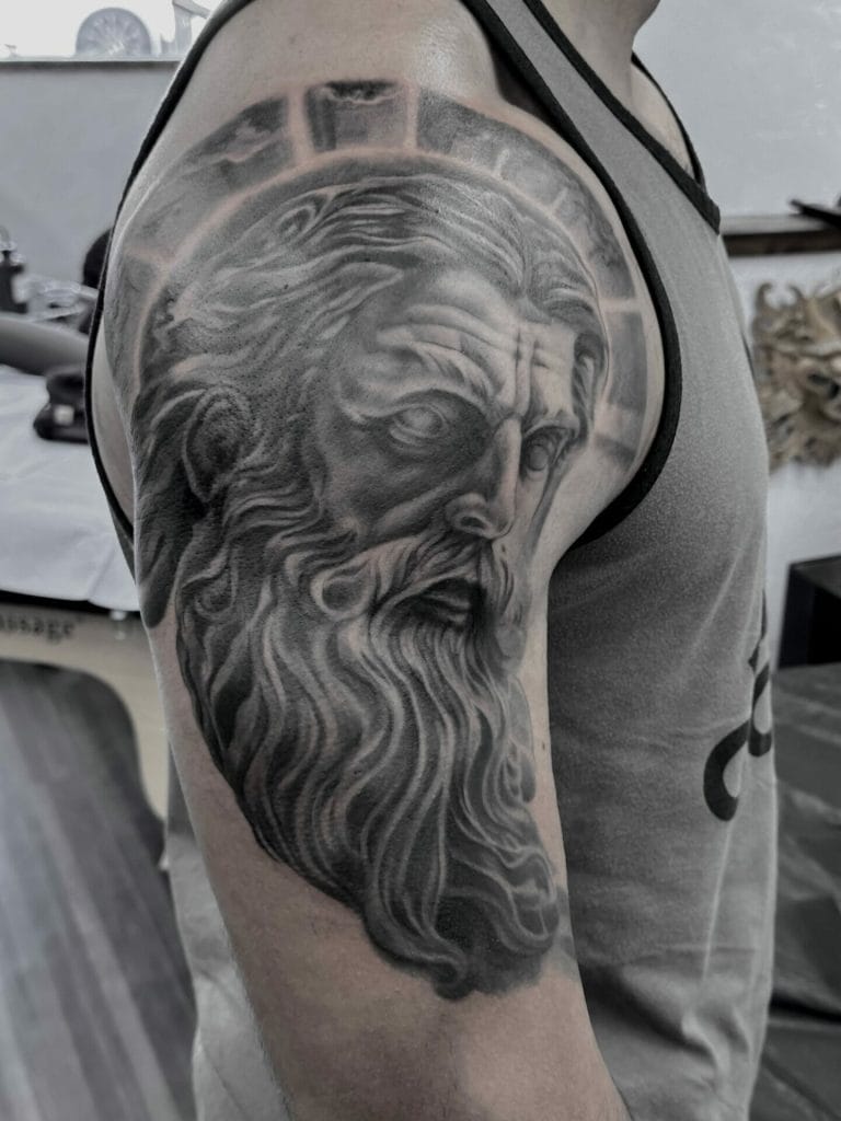 Detailed black and gray tattoo of a bearded mans face on upper arm.