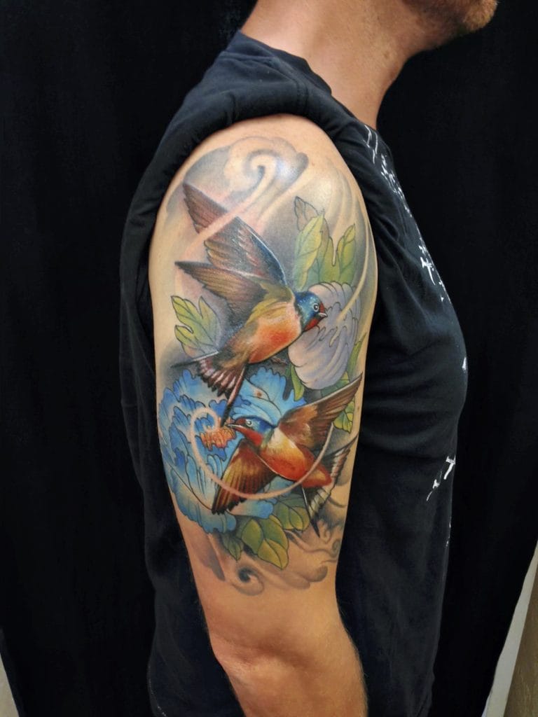 Colorful tattoo of two flying birds surrounded by leaves and clouds on persons upper arm.