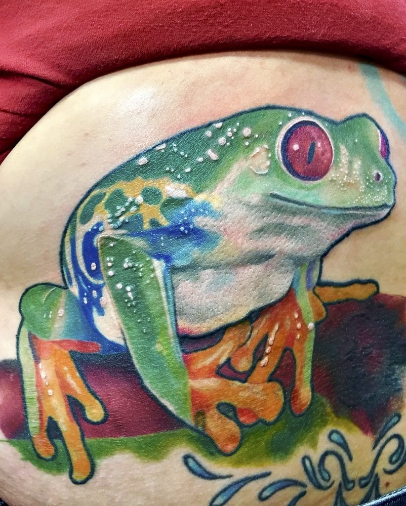 Colorful frog tattoo with large red eyes, sitting on a reddish surface.