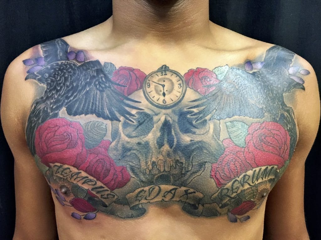 Tattoo of a skull with roses, ravens, a clock, and a banner reading Never Forget across the chest.