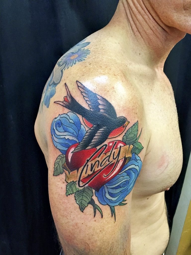 A tattoo of a swallow, red heart with Lindy script, and blue roses on a persons upper arm.