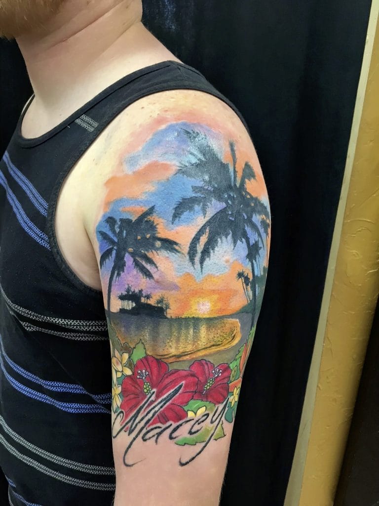 Colorful sunset over palm trees with hibiscus flowers and the word Aloha on the arm.