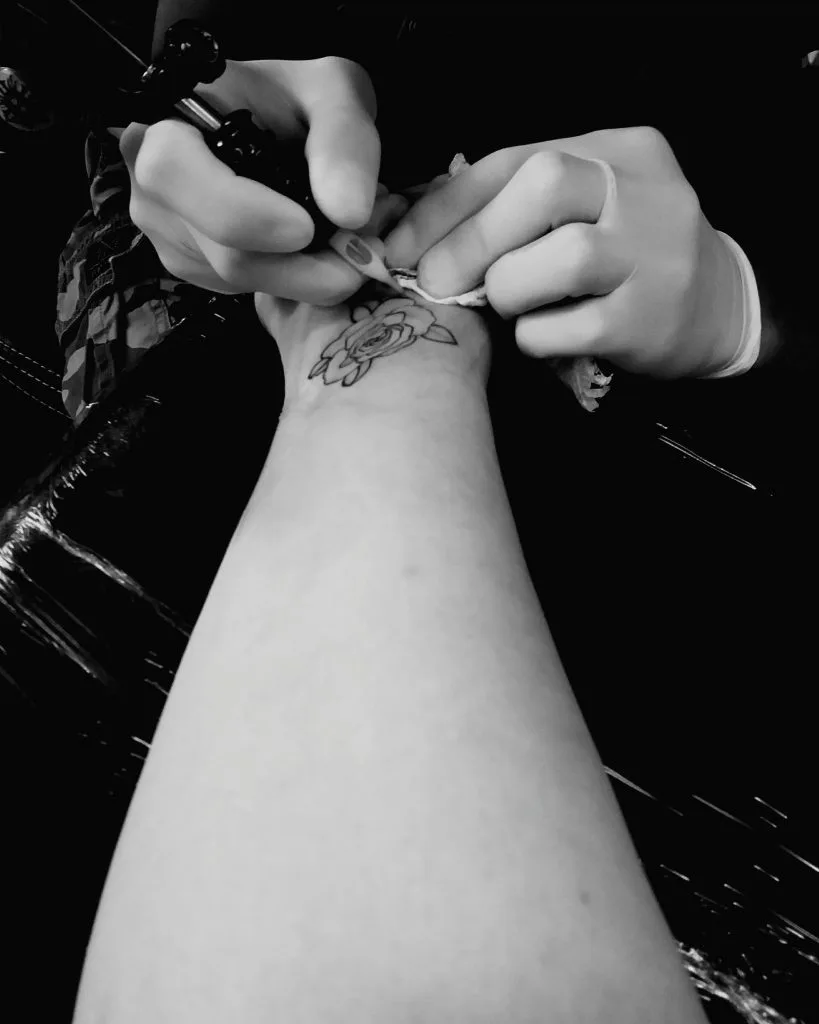 A tattoo artist wearing gloves is working on a floral tattoo with white ink highlights on a person's forearm, using a tattoo machine.