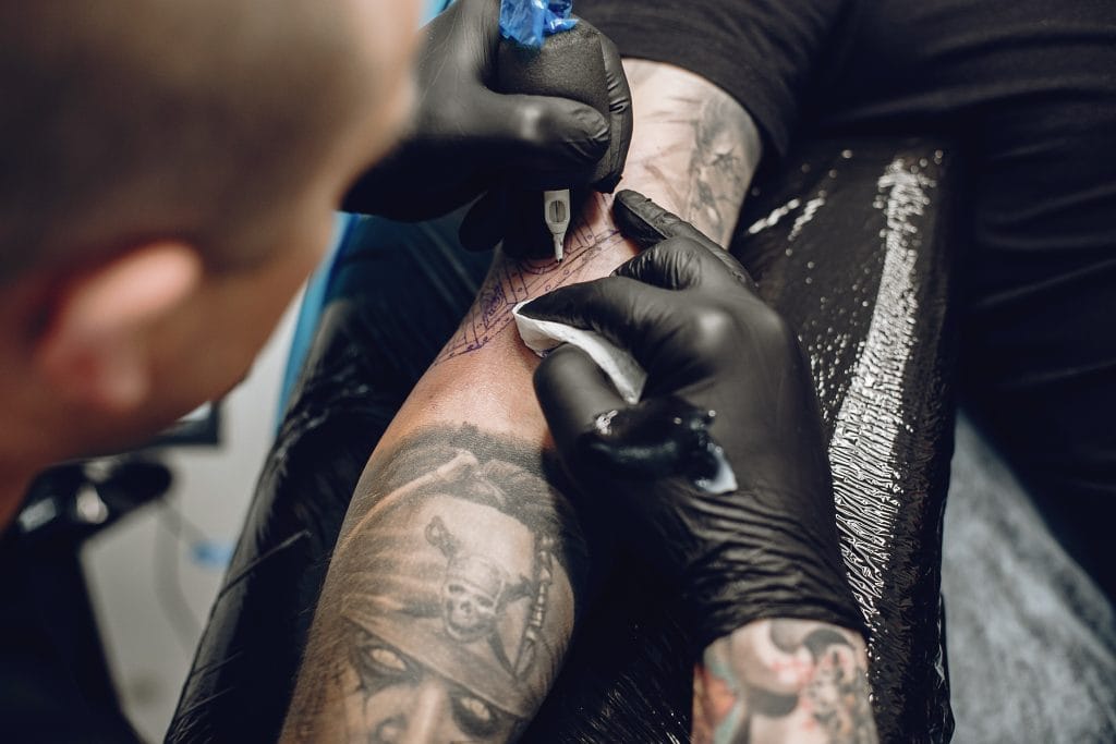 A tattoo artist wearing black gloves uses a tattoo machine to ink a design on a person's forearm, which already features a pirate skull and fresh white ink highlights.