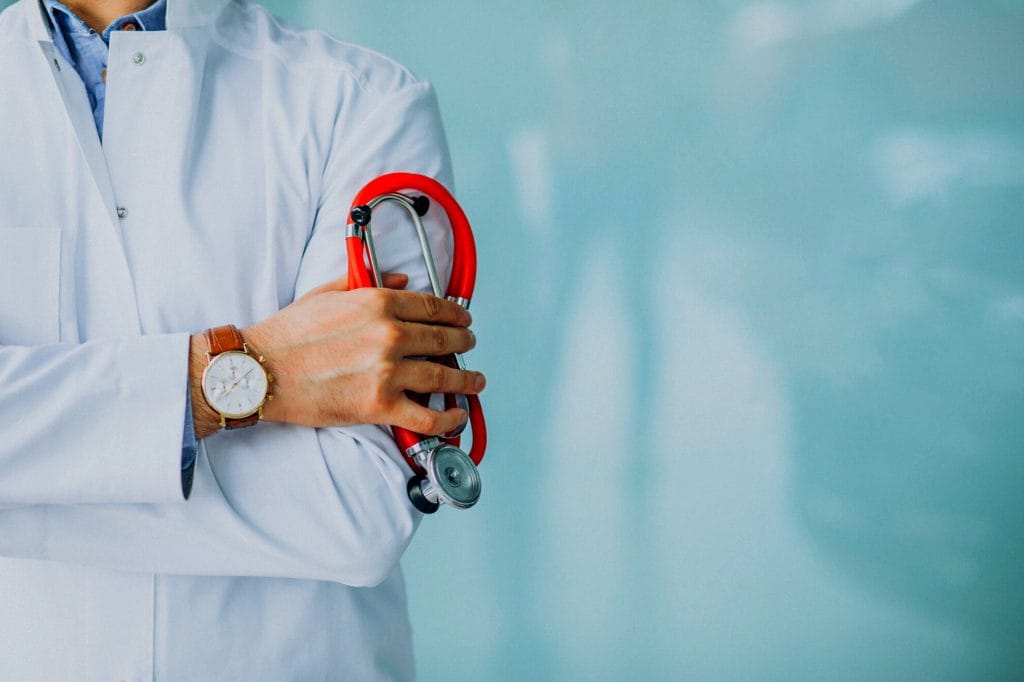 A doctor in a white lab coat, with tattoos visible on the forearm, holds a red stethoscope and crosses their arms, wearing a watch on the left wrist against a light blue background—ready for medical procedures.