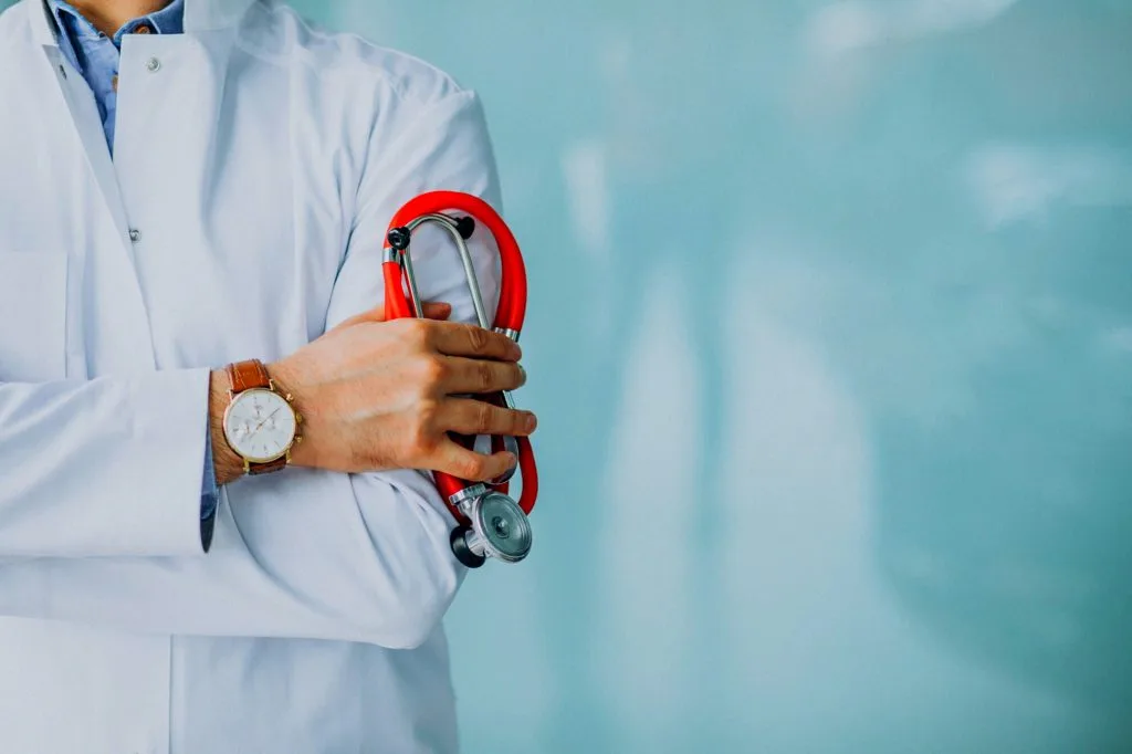 A doctor in a white lab coat, with tattoos visible on the forearm, holds a red stethoscope and crosses their arms, wearing a watch on the left wrist against a light blue background—ready for medical procedures.