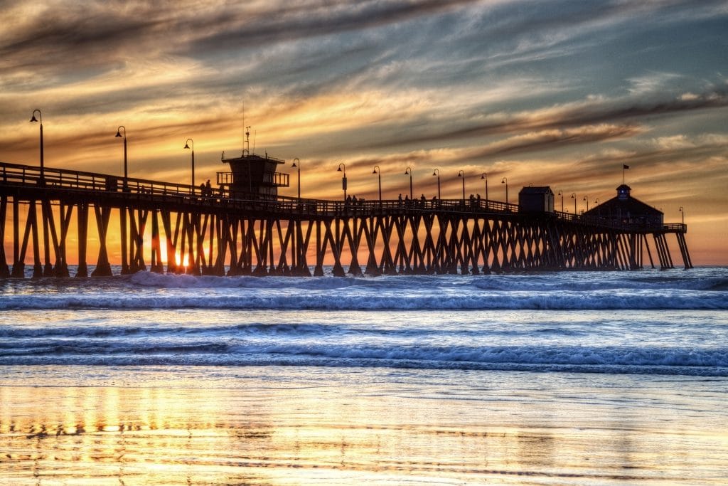 A long wooden pier extends over ocean waves at sunset, with dramatic clouds and silhouetted structures visible against the colorful sky—one of the iconic photo spots along San Diego beaches.