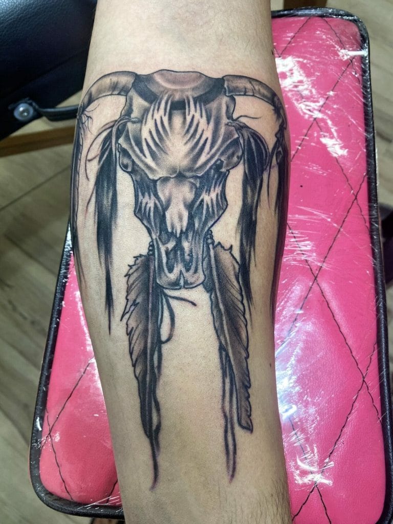 Black and gray tattoo of a skull with horns and feathers on a persons forearm.