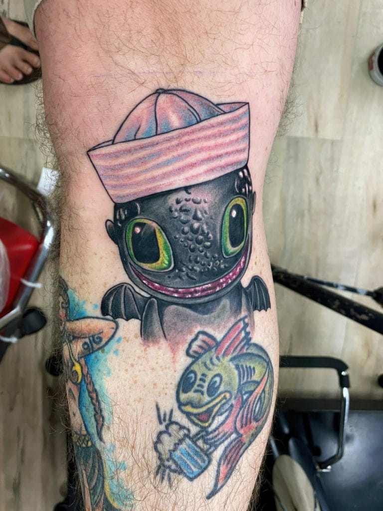 Tattoo of a cartoon dragon with large green eyes, wearing a pink striped hat, and holding a fish.