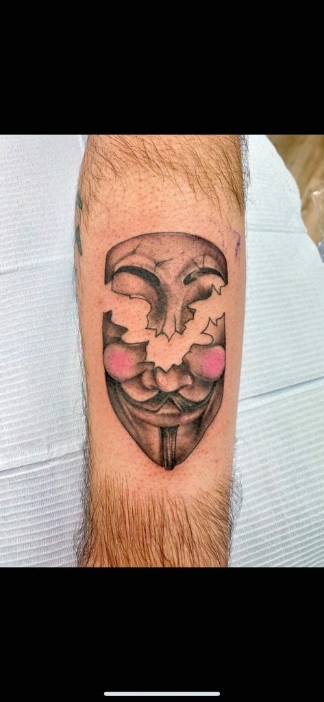 Tattoo of a stylized Guy Fawkes mask with pink cheeks on a persons arm.